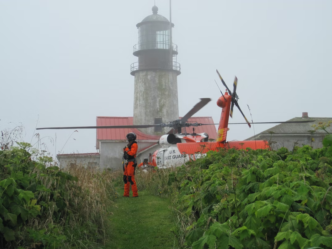 person in an orange jumpsuit uniform stands next to a coast guard helicopter. There is a lighthouse in the background.