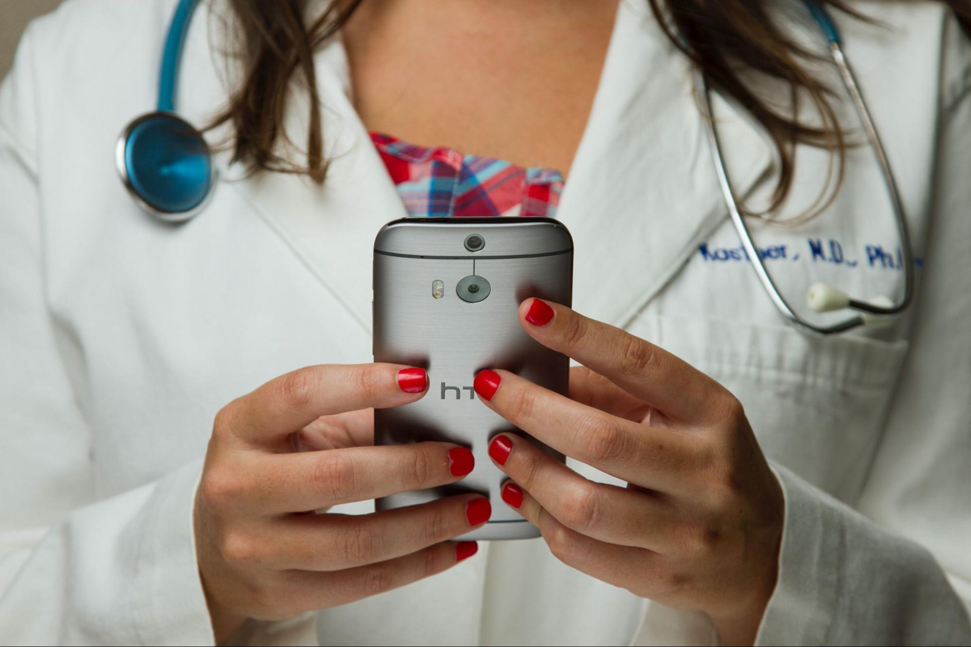 a doctor in a white lab coat wearing a stethoscope is using a cell phone to deliver telemedicine. Her fingernails are painted