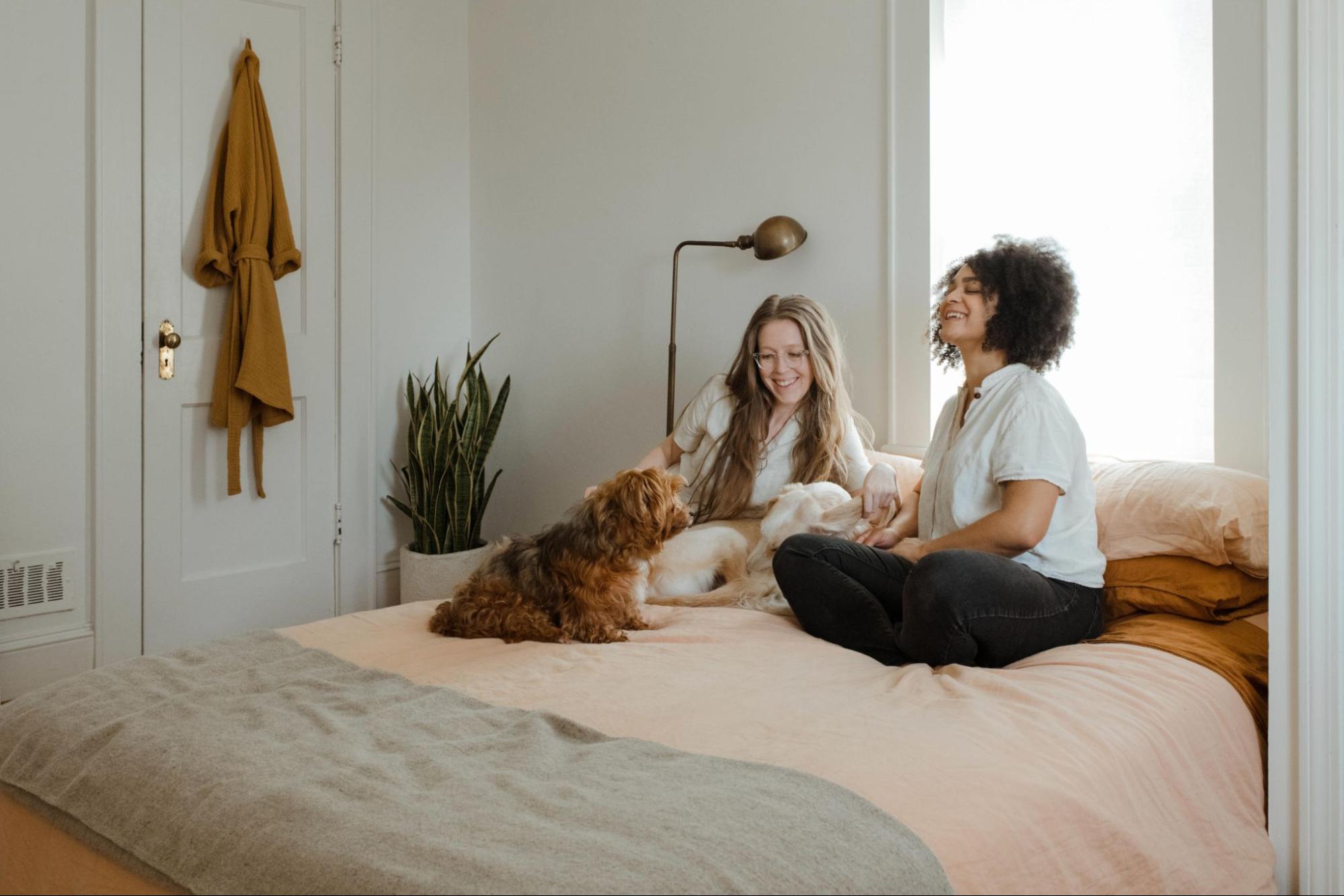Two female-presenting people sitting on a bed with their dogs. They are smiling.