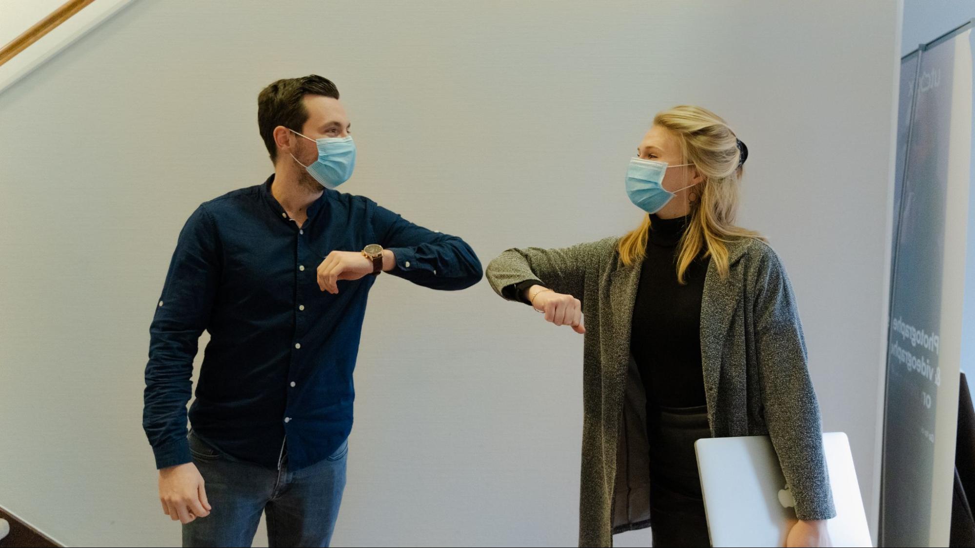Two people wearing masks associated with avoiding spread of COVID 19 are bumping elbows in greeting.