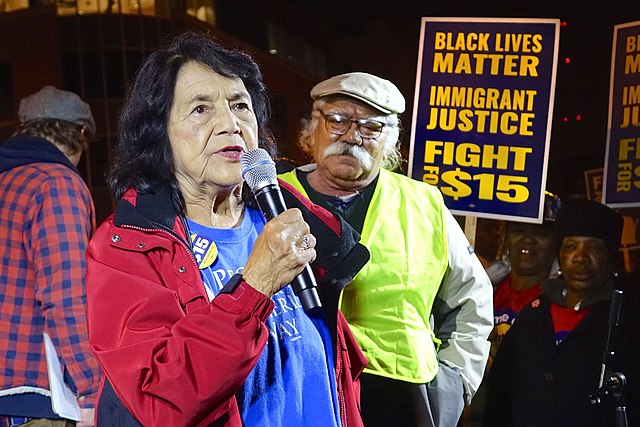 Activist Dolores Huerta is speaking into a microphone as people watch. A person holds a sign that reads, "Black lives matter, immigrant justice, fight for $15."