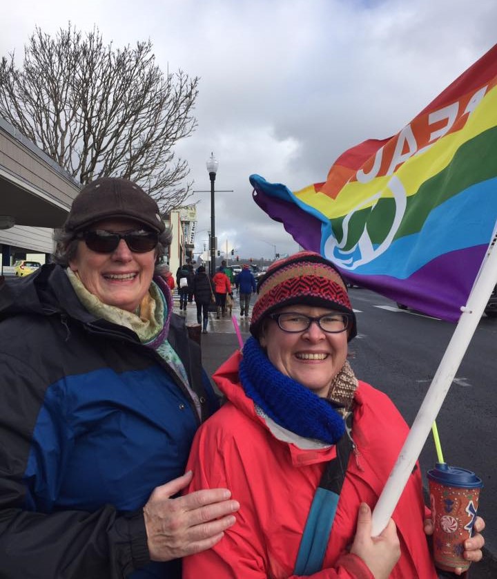 Two smiling women stand on the side of a street during the Women's March. One holds a rainbow flag that reads, "Peace" and holds a coffee.