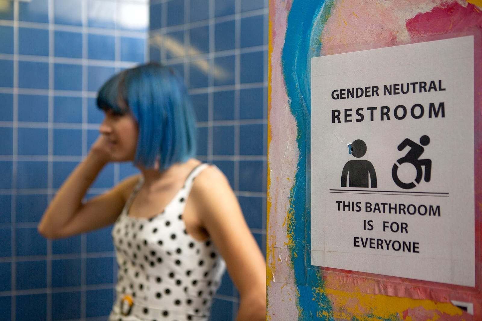 A female-presenting person stands in a restroom. A sign on the wall reads, "Gender neutral restroom, this bathroom is for everyone."