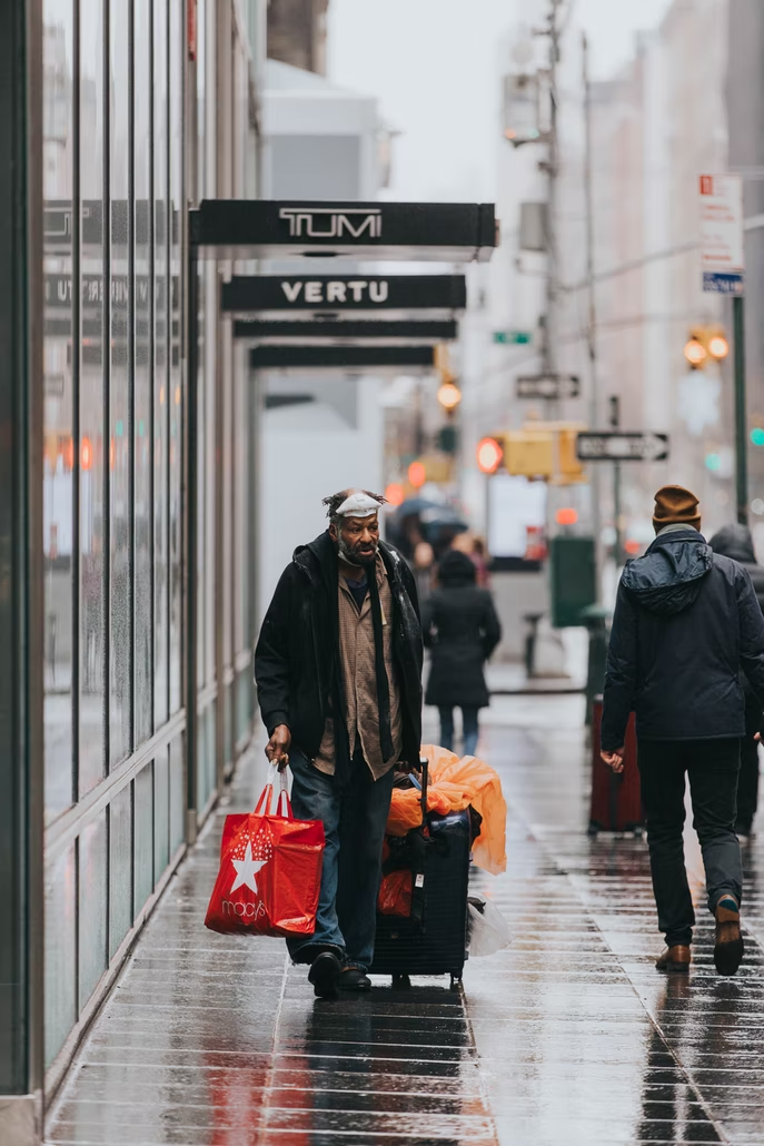 An older Black man walks down a rainy street rolling a large piece of luggage and a shopping bag containing his possessions. The surrounding stores sell high end luggage and other goods.