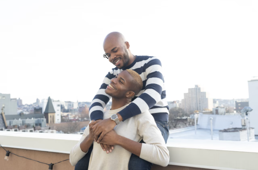 Two smiling, Black men are on a rooftop holding each other in an affectionate embrace.