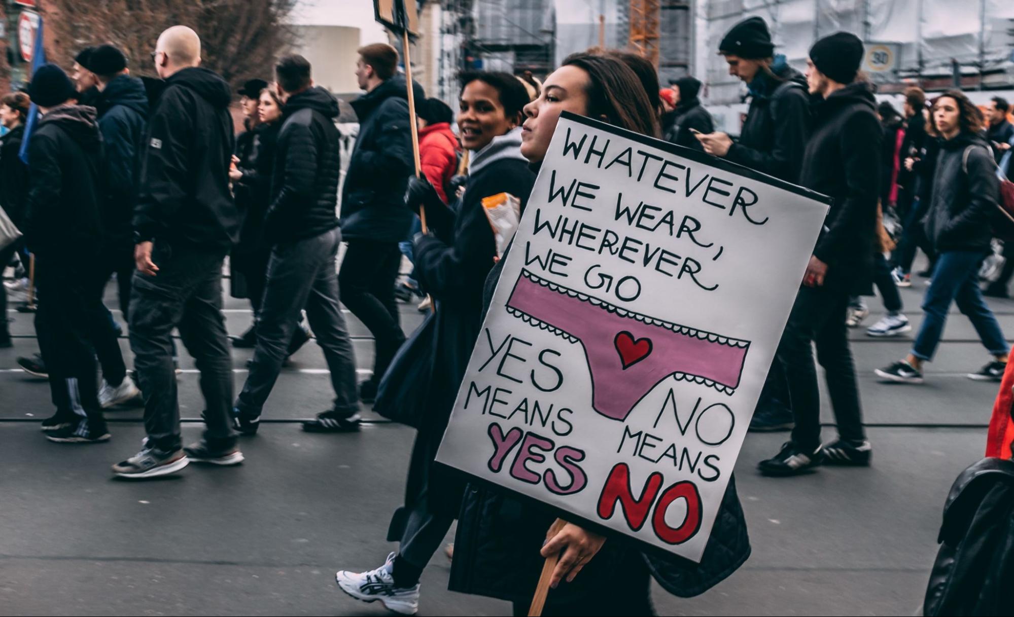 A crowd is walking down a street. One woman holds a sign that reads, " Whatever we wear, wherever we go, yes means yes and no means no."