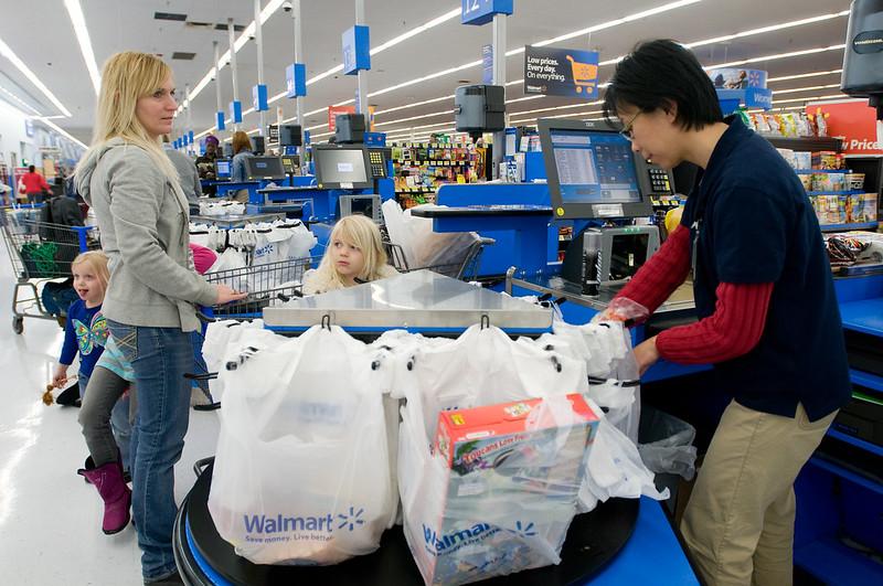 A woman with two small children stands by the checkout of a grocery store while a man with glasses bags groceries