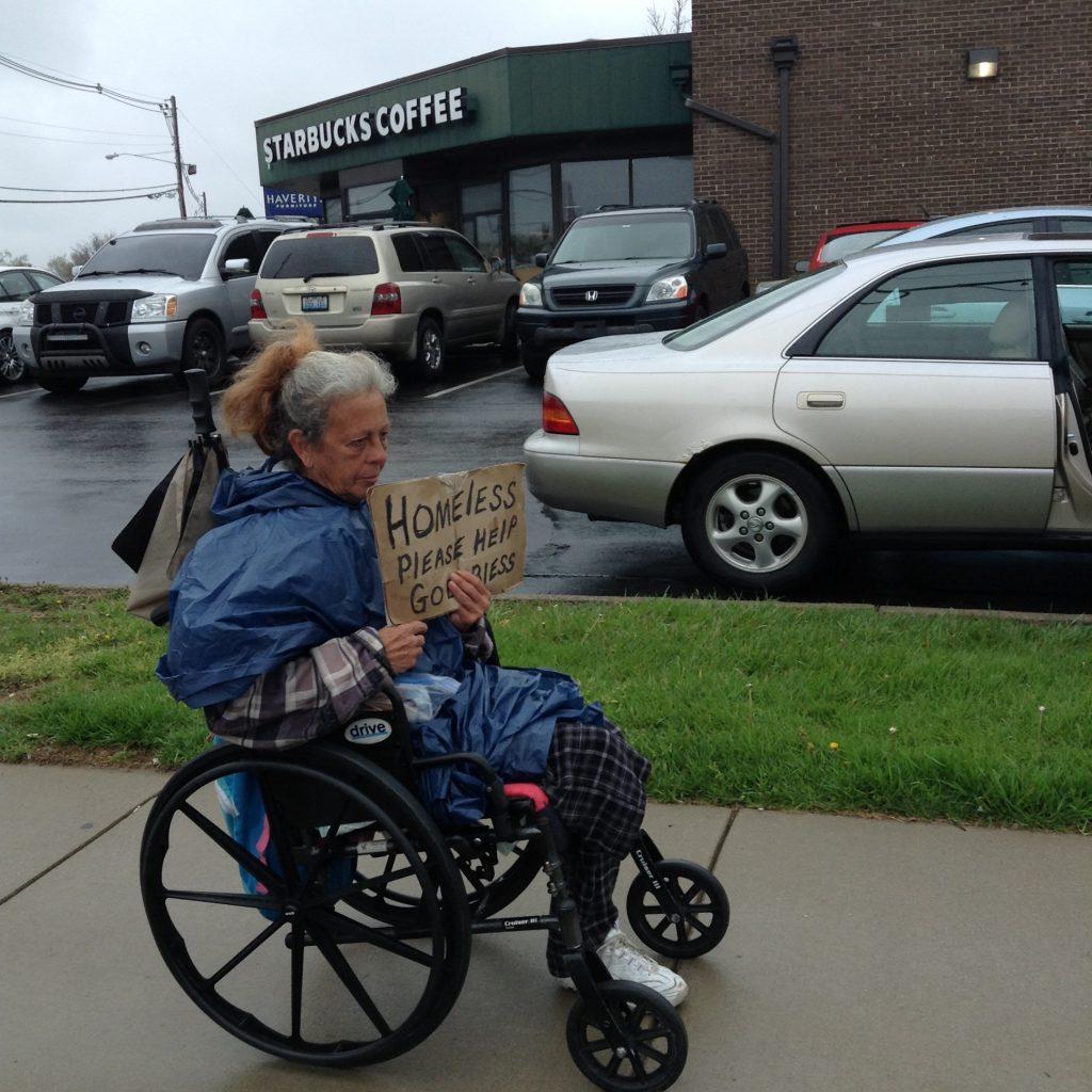 White woman in blue slicker sitting in wheelchair with sign "HOMELESS please help GOD bless"