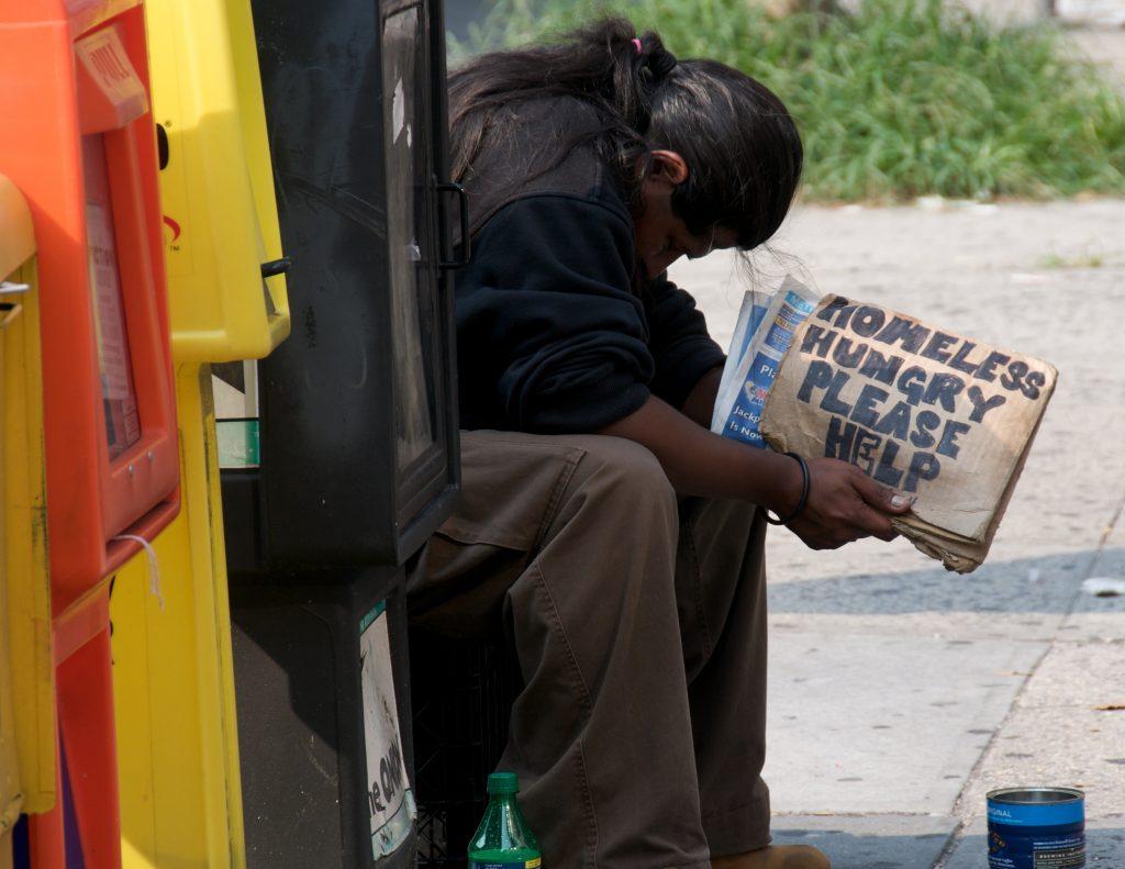 Person with ponytail sitting bowed over by newspaper boxes with sign "homeless hungry please help"