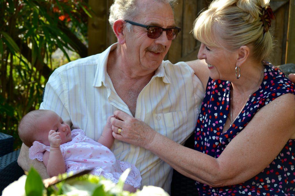 An elderly white man with sunglasses holding baby wearing pink and hugging a blonde white woman