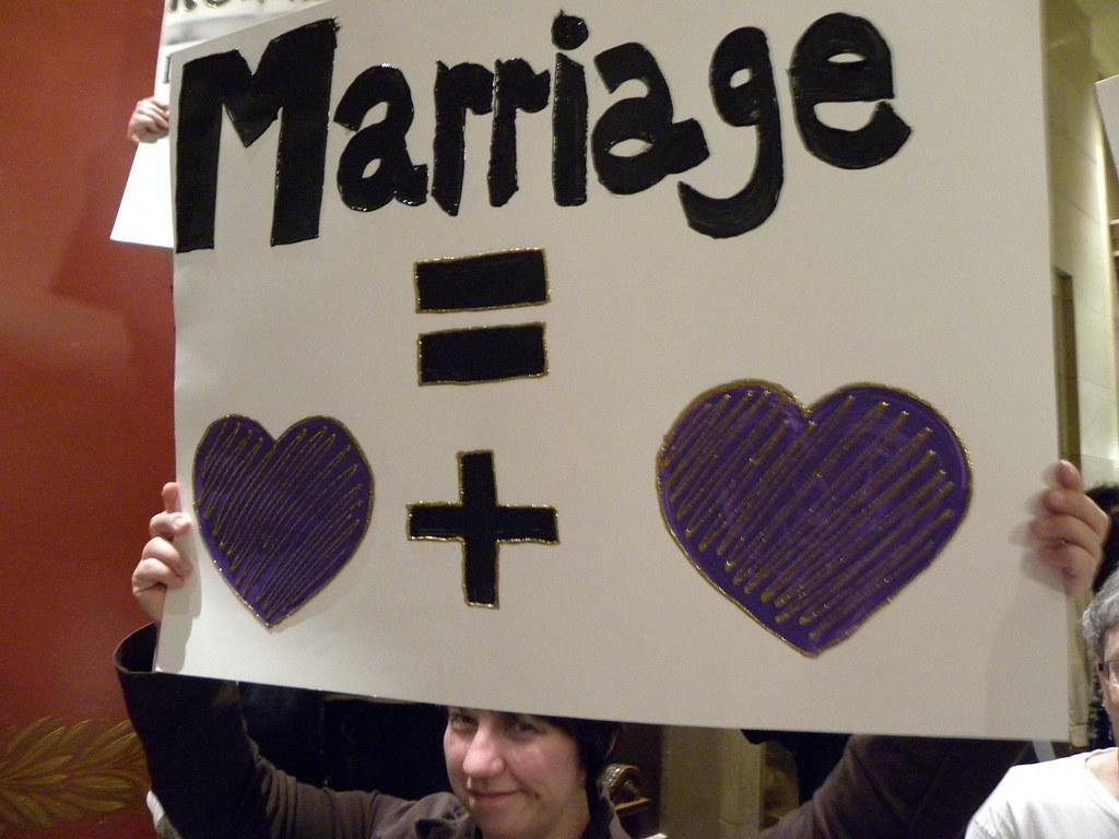 White woman holds sign "Marriage = a heart plus a heart