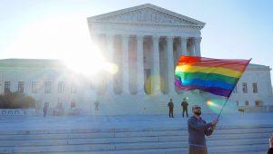 Photograph of a person waving a rainbow colored flag in front of the Supreme Court of the USA.
