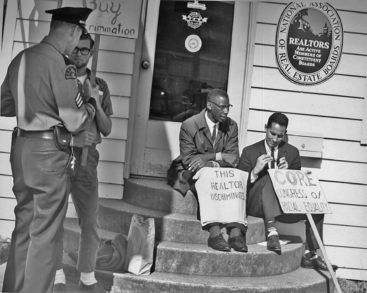 One law officer and three male protesters in front of the National Association of Real Estate Boards office. Signs say &quot;This Realtor Discriminates&quot; and &quot;Core Congress of Racial &lt;span class=&#039;glossary-term&#039;&gt;Equality&lt;/span&gt;&quot;.
