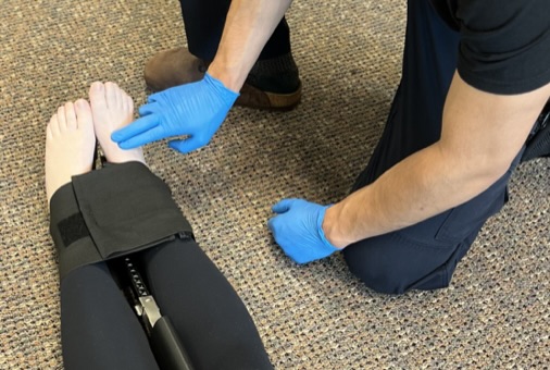 Male with gloved hands, placing his pointer and middle finger on the dorsal aspect of a supine patient's to locate dorsalis pedal pulse. Patient has traction splint secured.