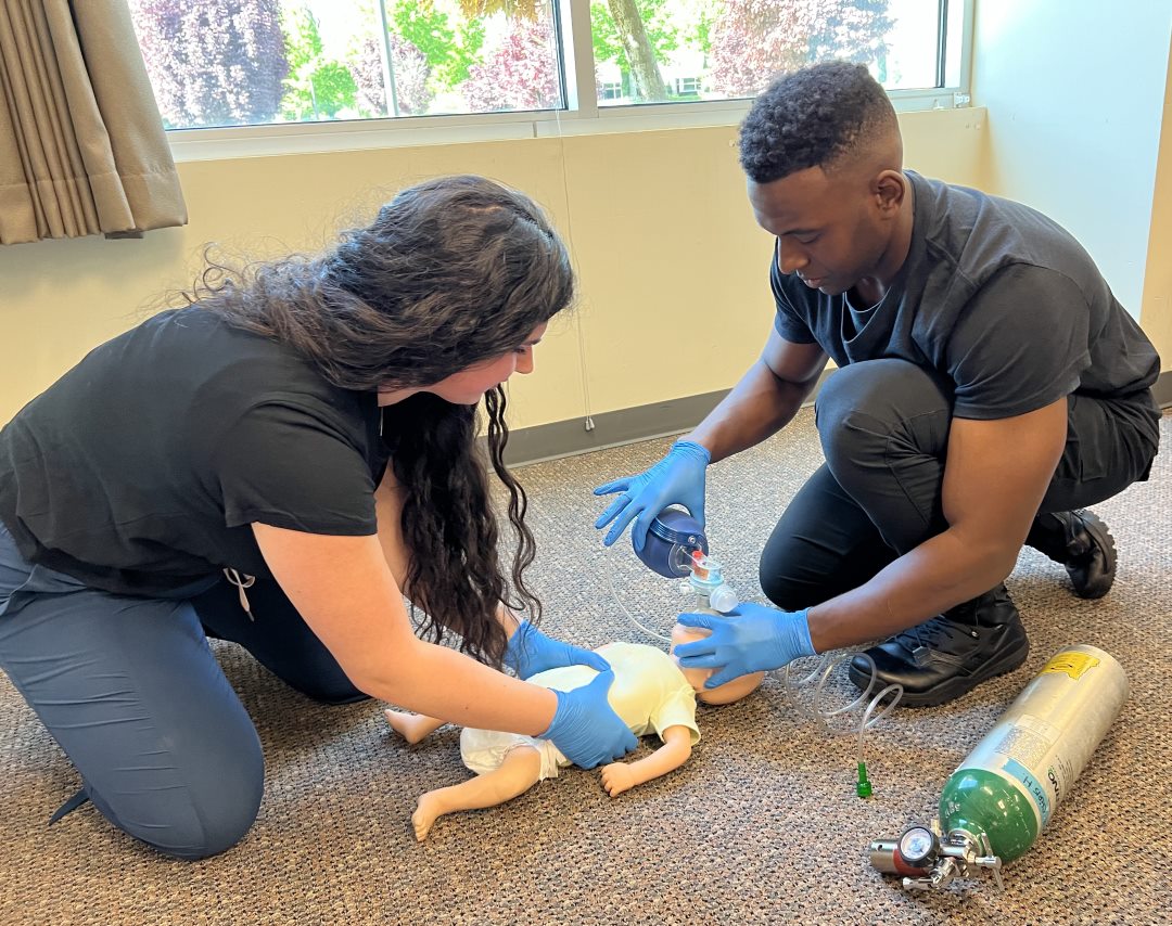 Two gloved EMTs performing 2 person infant CPR on infant manikin.
