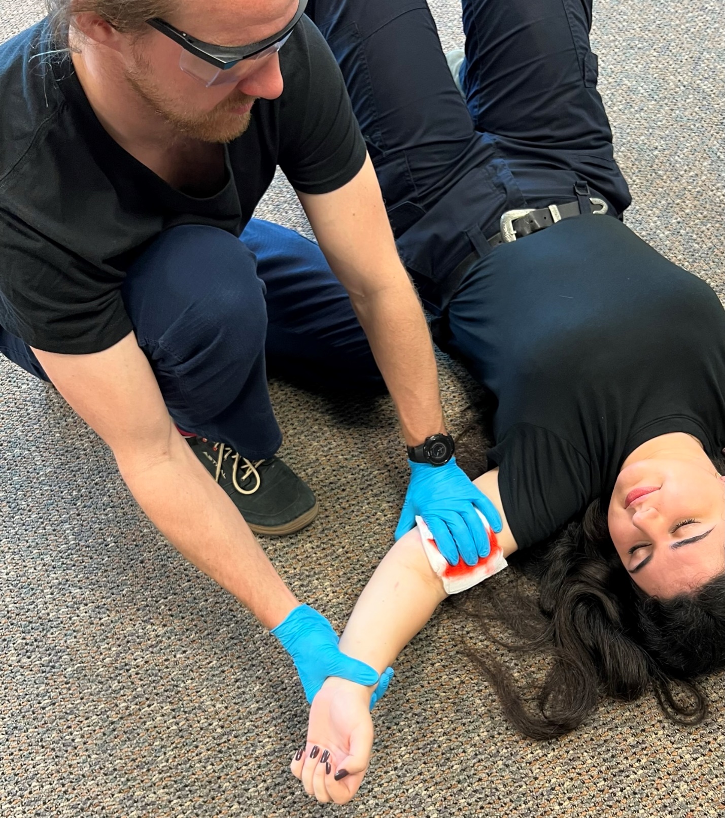 Photo of gloved EMT kneeling next to supine patient while holding direct pressure with blood tinged gauze to patient's left upper arm.