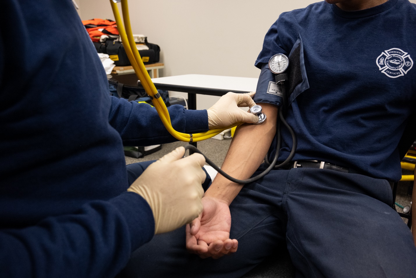 A gloved person, obtaining a blood pressure by auscultation on a patient. The bell of the stethoscope is on anterior/medial portion of the patients' arm.