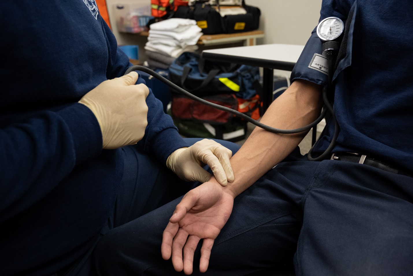 A gloved EMT obtains blood pressure by palpation using two fingers on a patient's wrist.