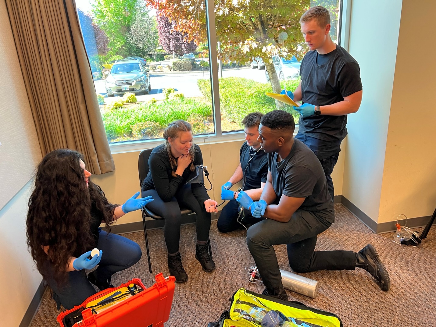 A group of 4 EMS providers, knelt down around patient that is seated in chair. One of the providers appears to be asking question with uplifted hand and slight should shrug while looking at another responder.