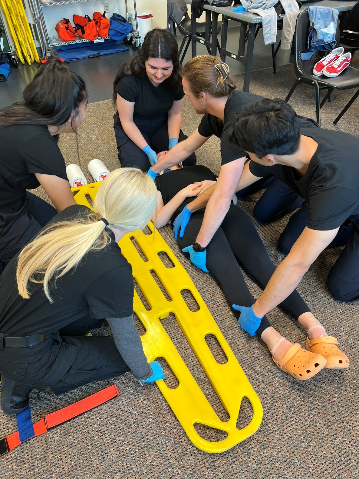 Photo of 6 gloved EMTs. Two EMTs are preparing to logroll supine patient with an EMT at the patient's head holding c-spine stabilization. The two remaining EMTs are preparing to slide backboard under patient.