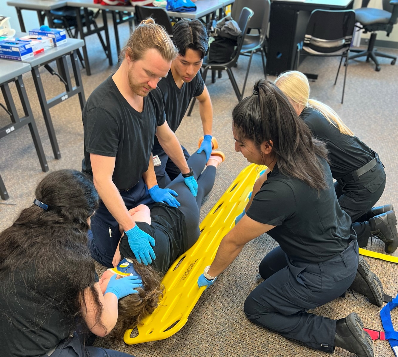 Photo of 5 gloved EMT's. Two EMT's have logrolled the patient, an EMT is holding manual c-spine stabilization while the remaining two EMT's are sliding backboard under patient.