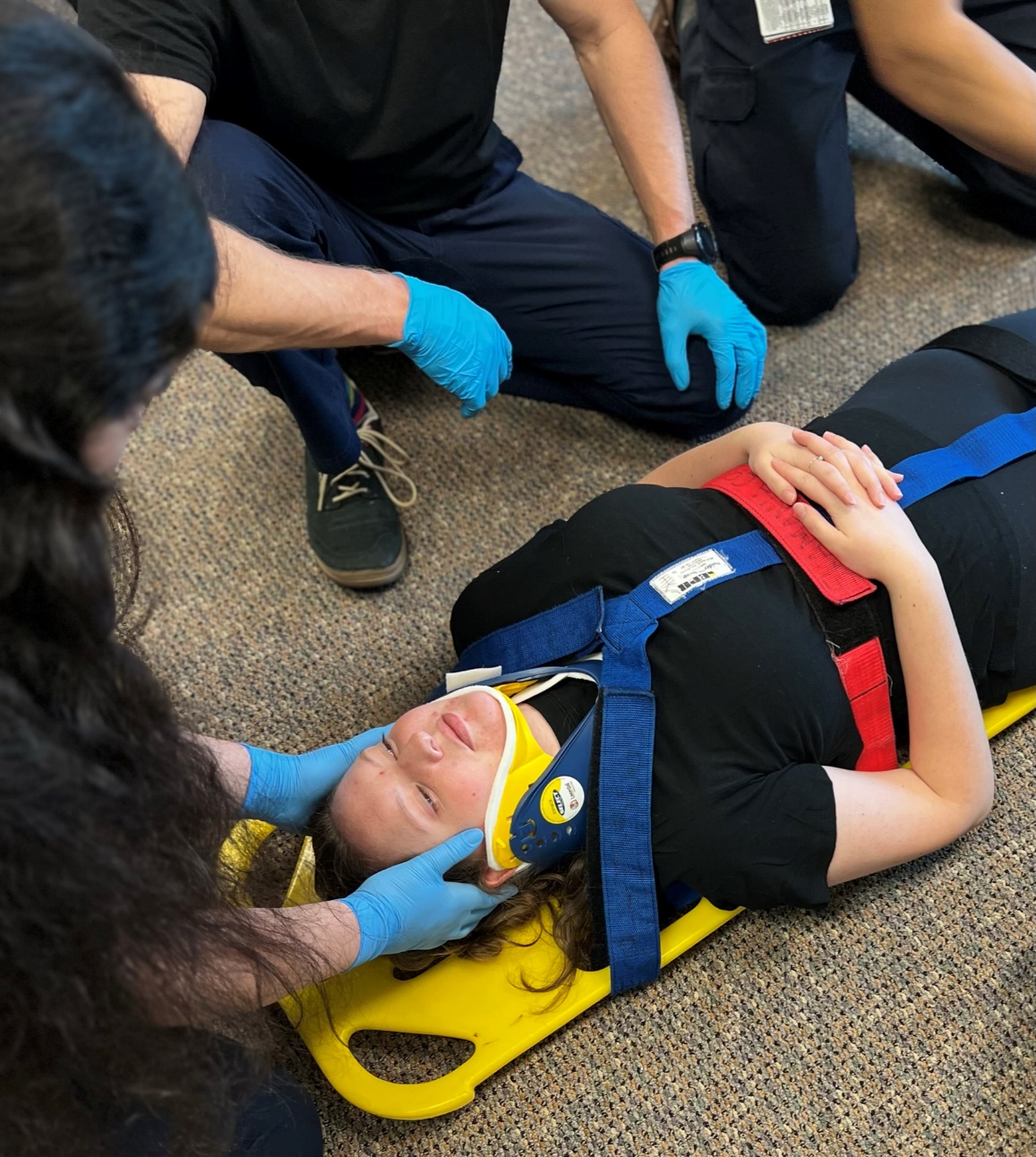 Photo of patient secured onto backboard with straps with gloved EMT holding manual C-spine immobilization.
