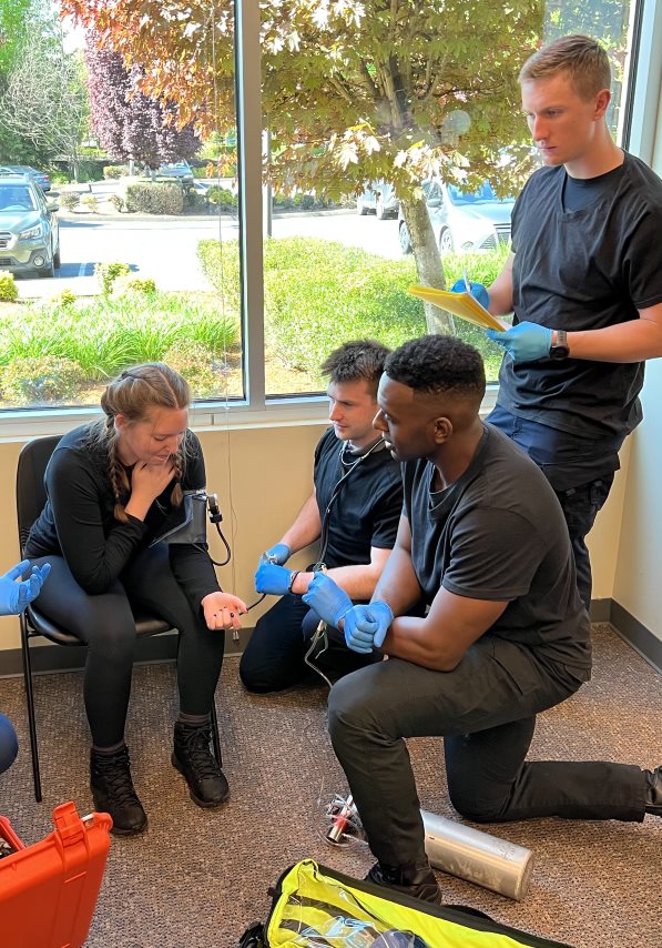 Photo of team dynamics. One EMT obtaining vital signs, another interviewing the seated patient, a third EMT documenting information.