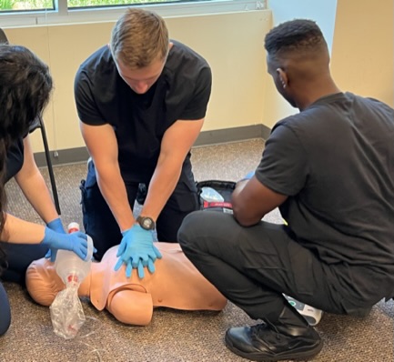 Photo of 3 gloved EMTs. One EMT is performing chest compressions on manikin, another EMT is providing ventilations via BVM and the third EMT is preparing AED.