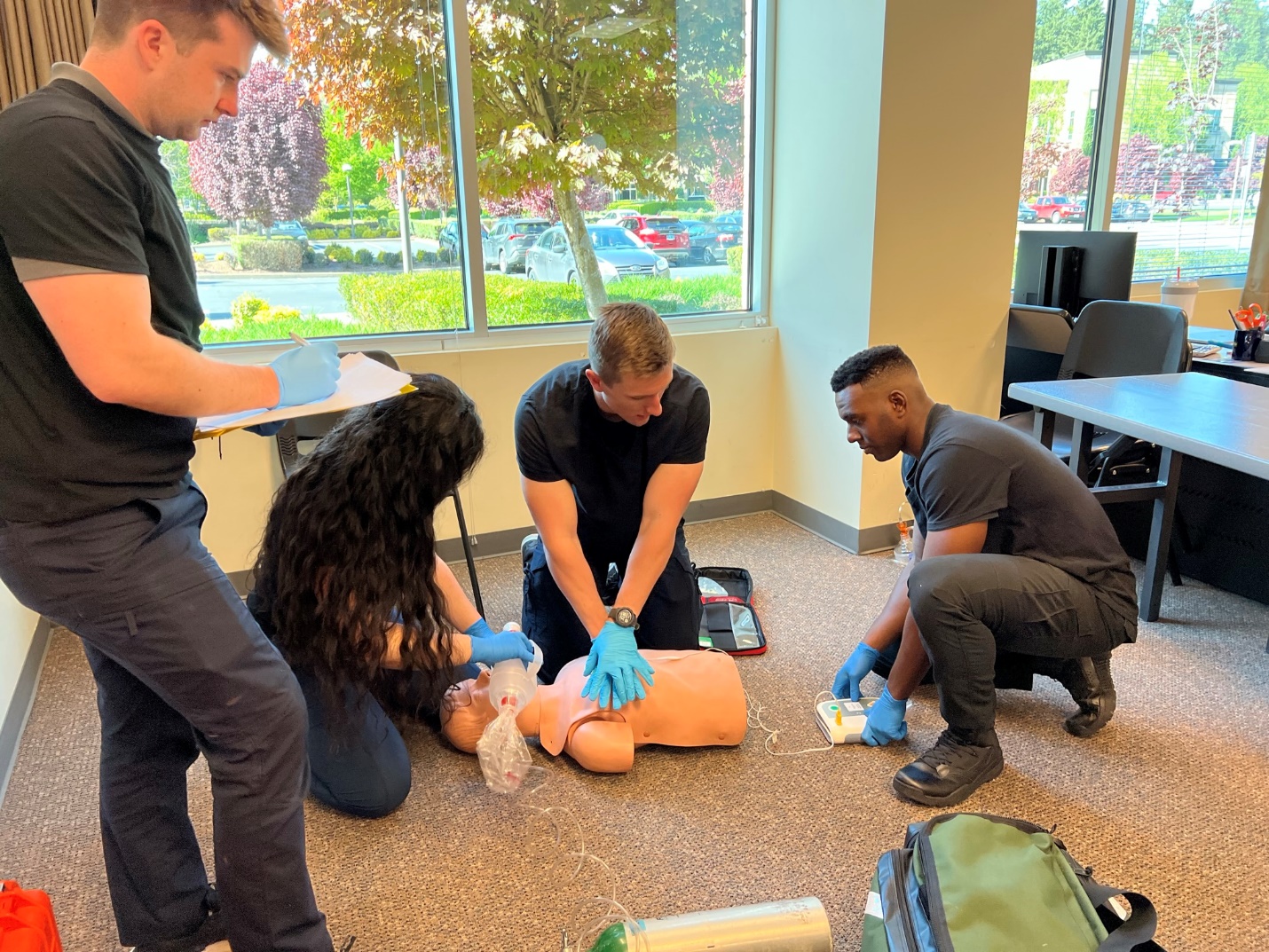 Photo of 4 EMTs showing team dynamics. One EMT is performing chest compressions on manikin, another is providing ventilations via BVM, another is operating AED and the forth is documenting events