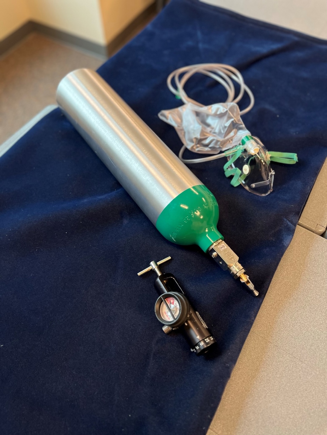 A photo of the equipment needed to deliver oxygen to a patient in the field via a non-rebreather mask, including a portable oxygen tank, an oxygen regulator, and an non-rebreather mask.