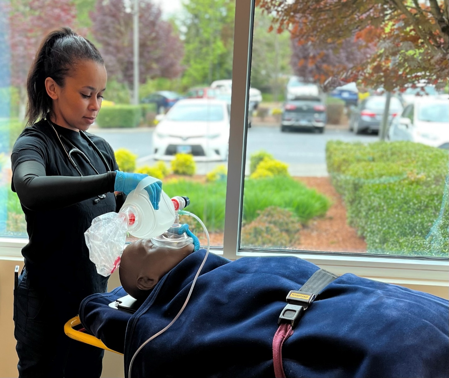 Gloved EMT at head of manikin that is on stretcher. EMT is ventilating the manikin with BVM with supplemental O2.