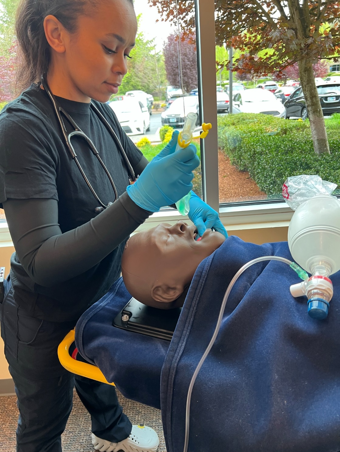 Gloved EMS clinician standing at foot of manikin using scissor technique to open manikin's airway with i-Gel in other hand, preparing to insert i-Gel.