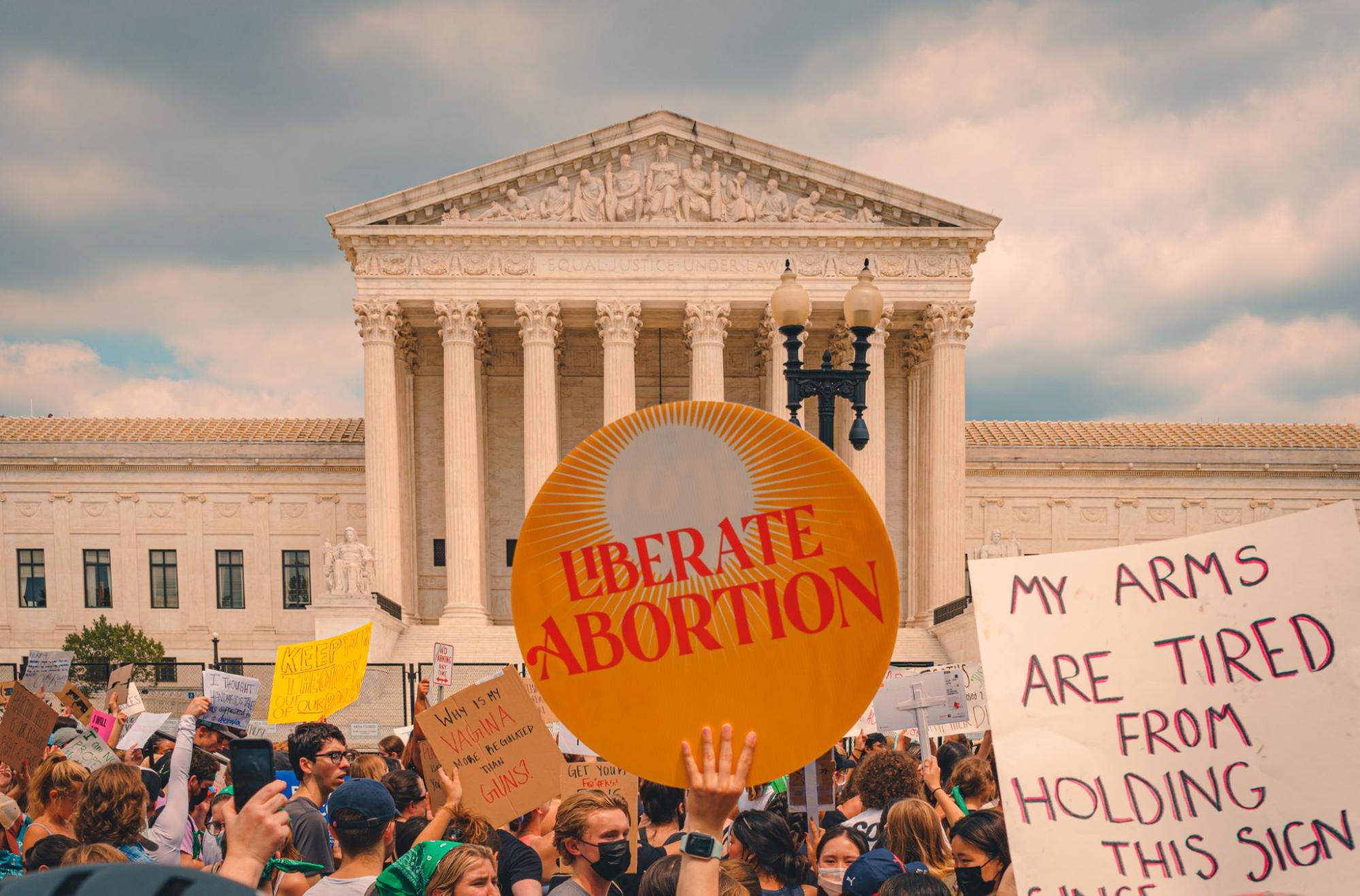 A crowd stands in front of the Supreme Court with signs that say liberate abortion and my arms are tired from holding this sign.