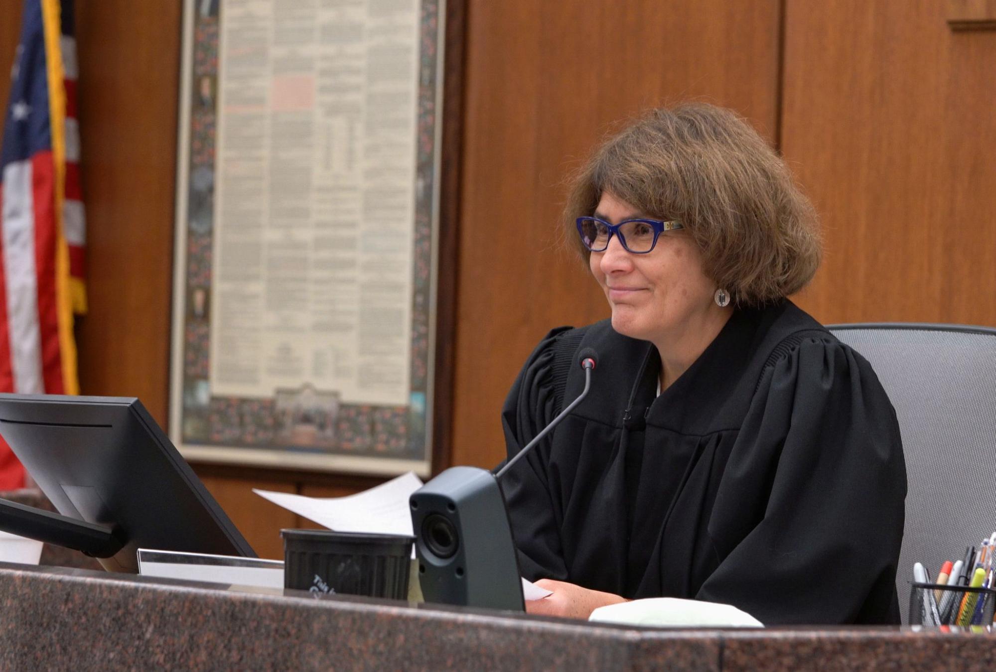 Judge Reding is smiling and has blue glasses, earrings, and brown hair.