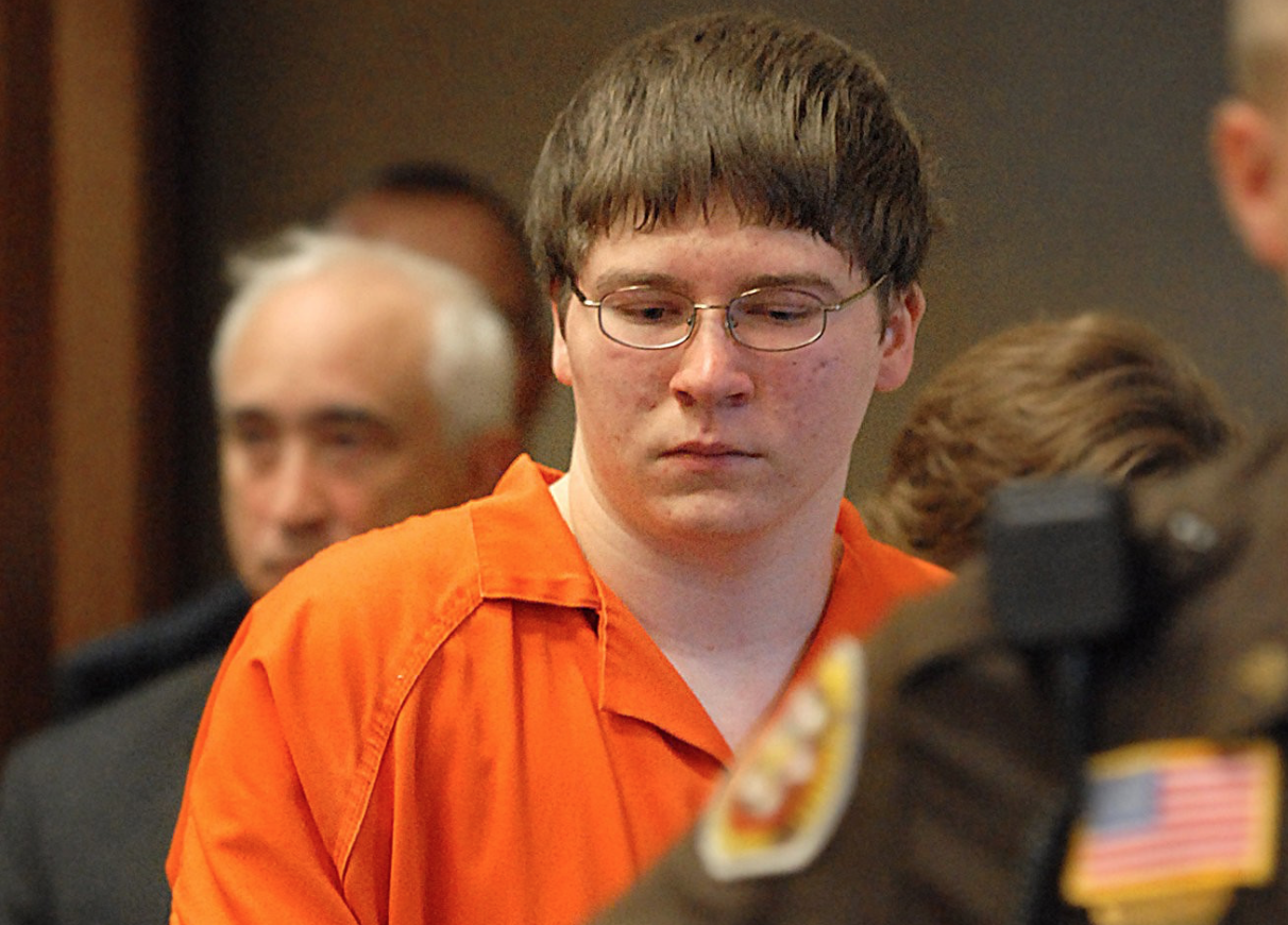 An image of Brendan Dassey, in the courtroom, wearing an orange jumpsuit with an officer and others people in the fore and background.