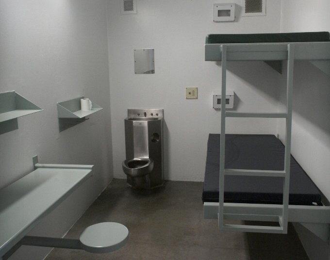 A photo of a facility cell showing a metal bunk bed with two mattresses, a toilet/sink combo, a mirror, two shelves, a metal table and stool.