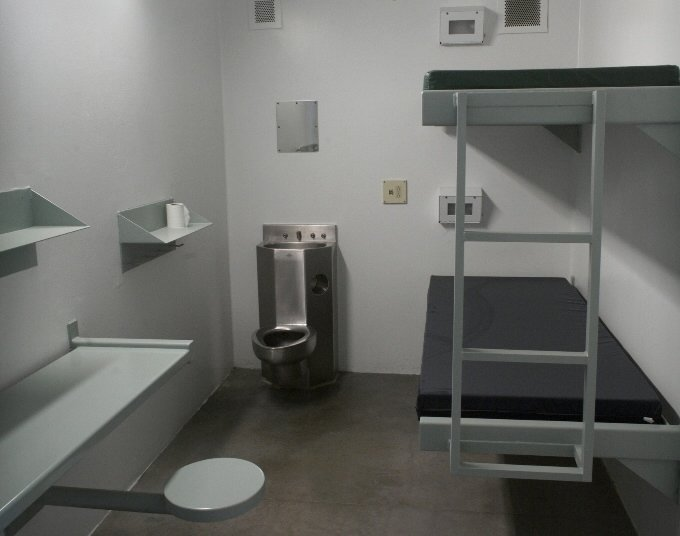 A photo of a facility cell showing a metal bunk bed with two mattresses, a toilet/sink combo, a mirror, two shelves, a metal table and stool. 