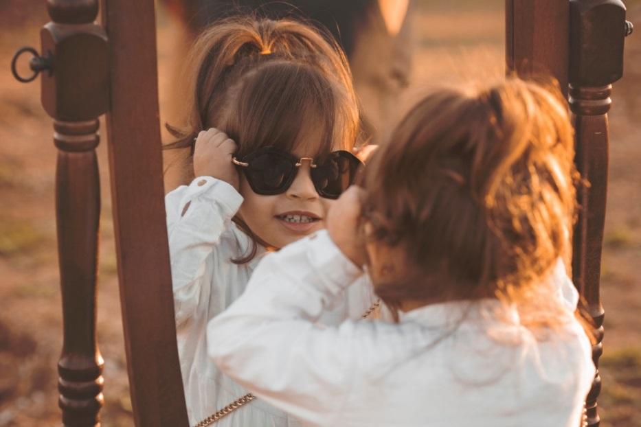 child wearing sunglasses and looking at her reflection in a mirror