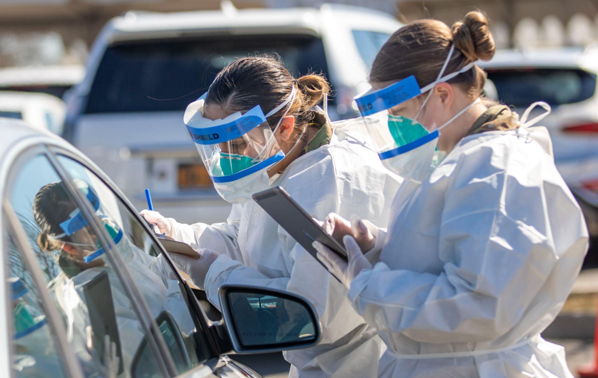 two women in professional grade covid protection stand in front of a vehicle taking notes.