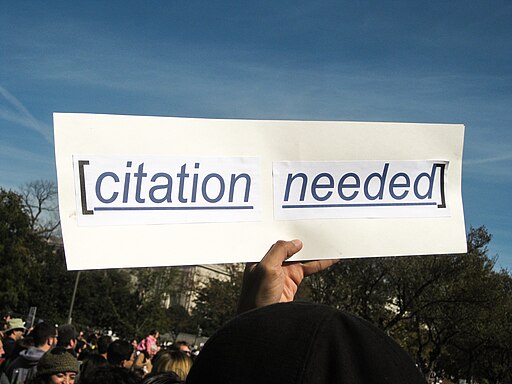 A hand with dark skin holds a handmade protest sign which reads "citation needed."