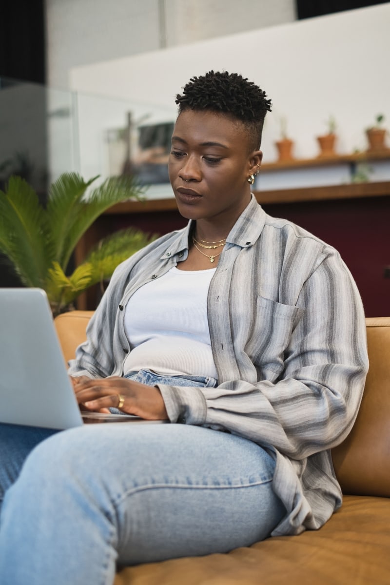 A dark skinned person sitting alone with a laptop, looking discouraged.