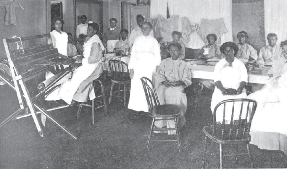 15 women at Central State Hospital staring at the camera. One sits at a loom and the rest sit at a worktable. Knitted or crocheted items hang on the wall behind them.