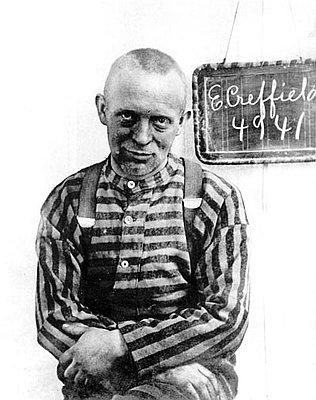 Edmund Creffield has light skin and a shaved head and stares at the camera while wearing a black and white striped prison shirt and suspenders. A sign hangs behind him that reads "E Creffield 4941"