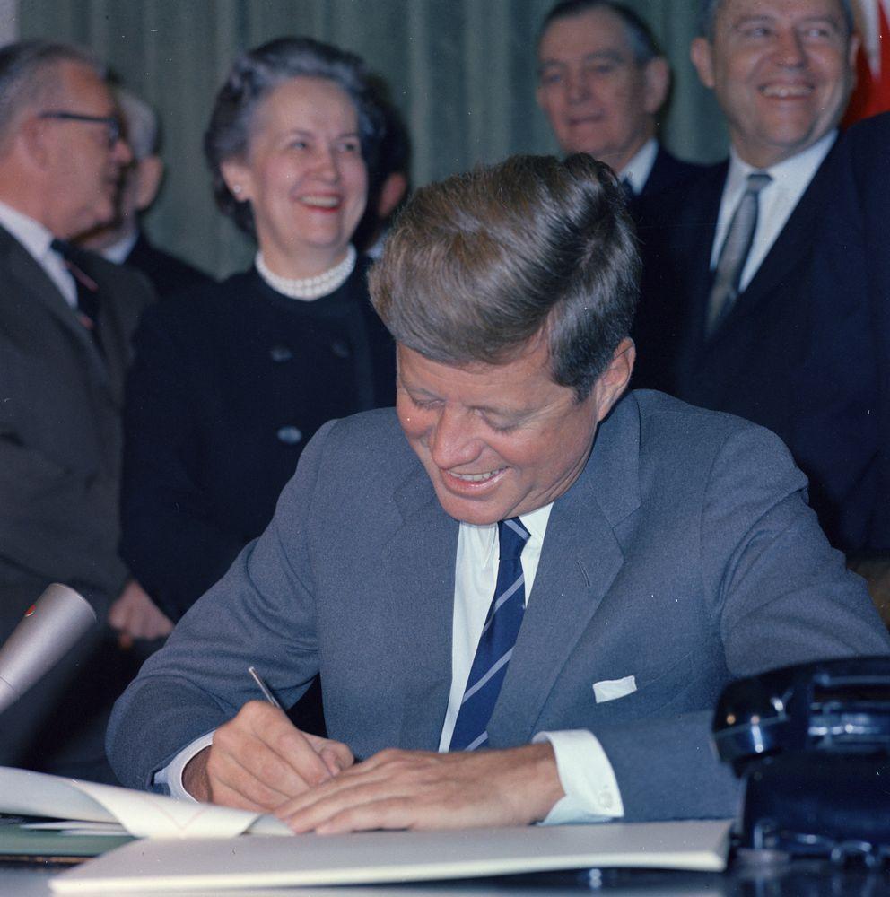 John F Kennedy smiles while signing the Community Mental Health Act into law. Other people smile behind him.
