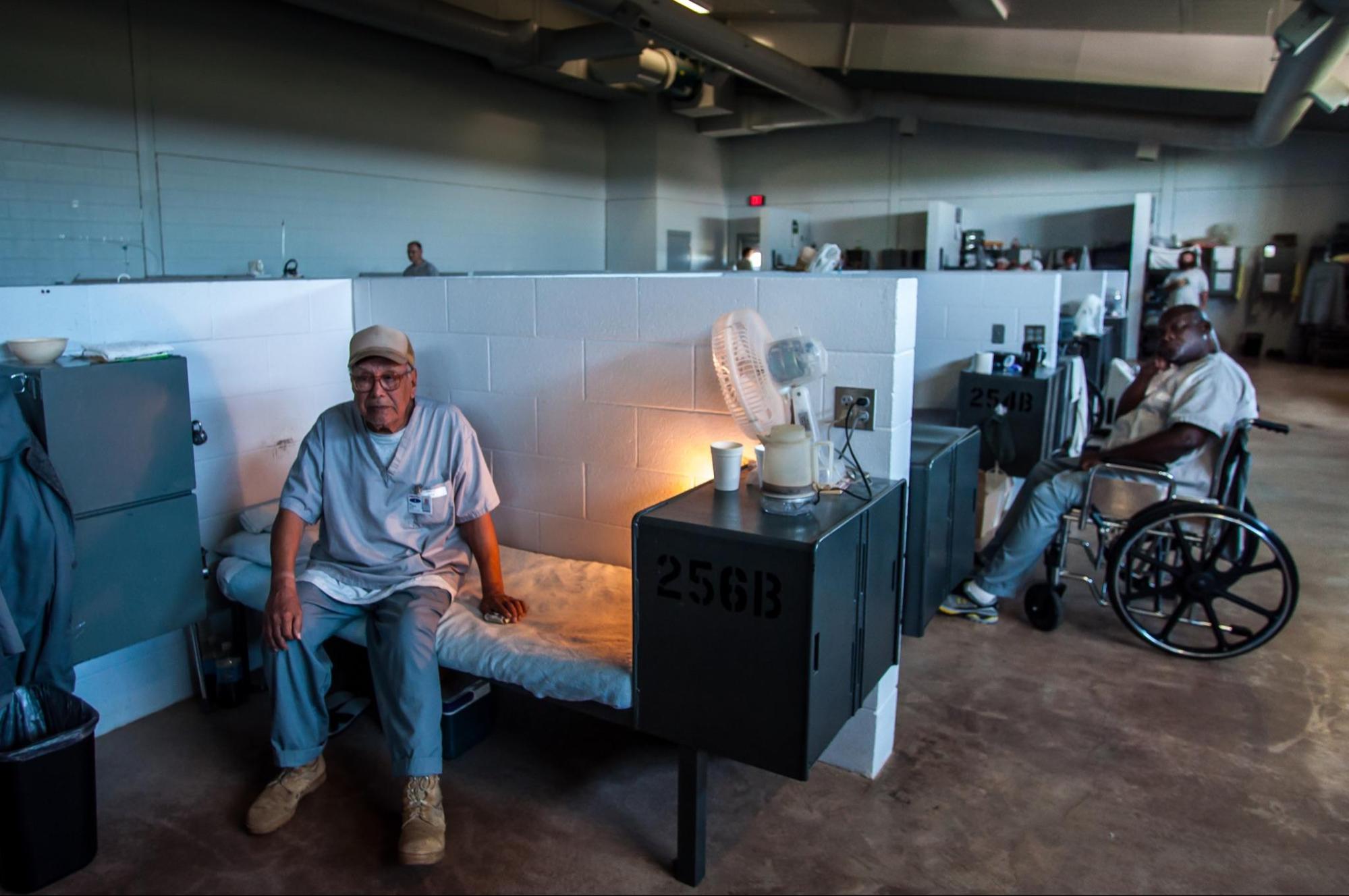 Male inmates at a prison. One sits on a bed and the other sits in a wheelchair.