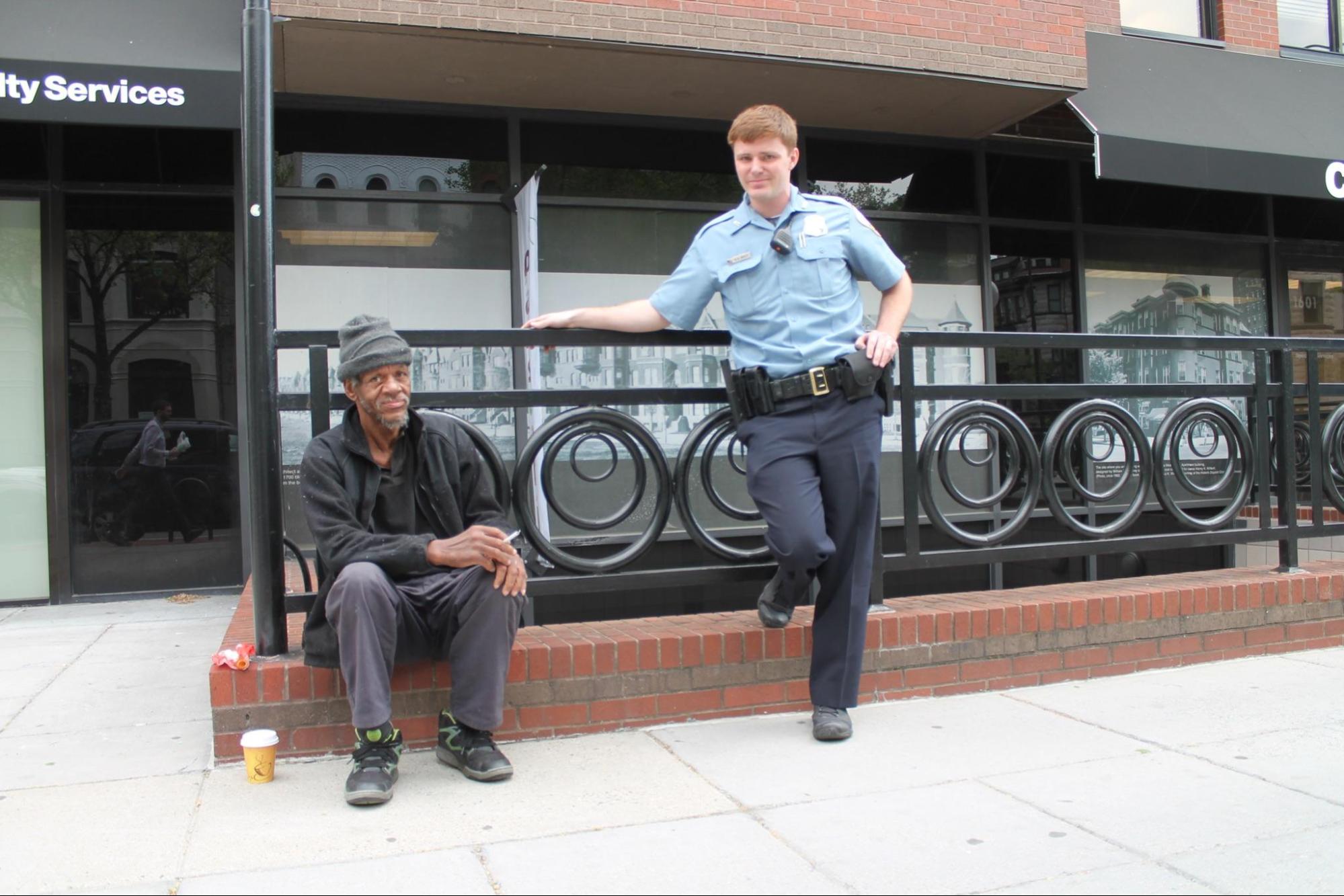 A young police officer standing next to an older man sitting on the sidewalk.