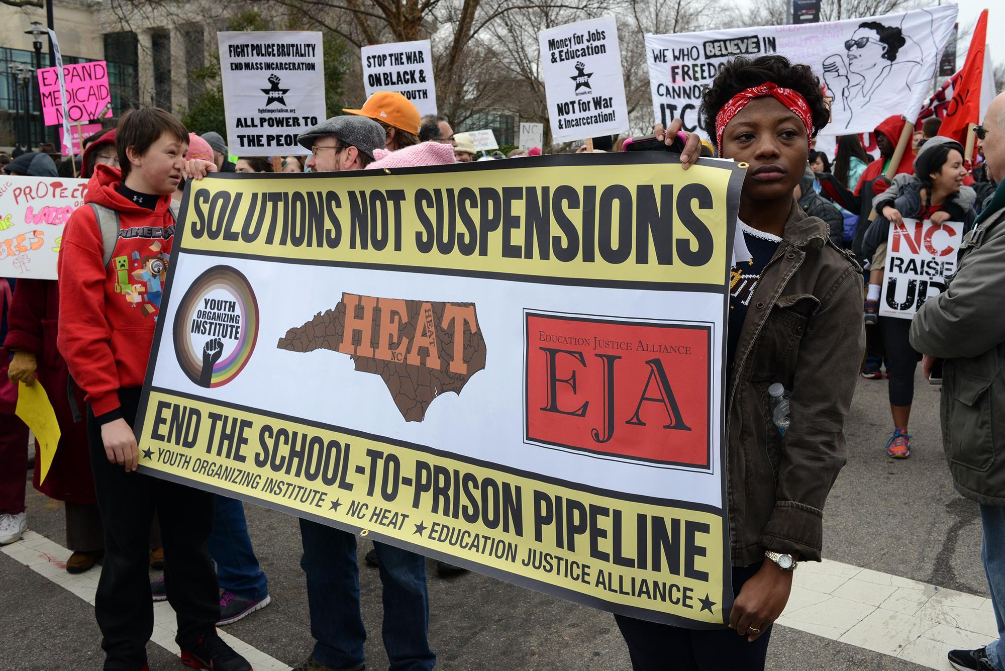 People at a protest holding a sign that reads "Solutions not suspensions. End the school to prison pipeline."