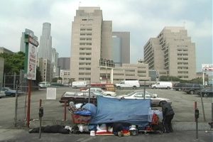 in the foreground we see a houseless living area with a tarp covering and a shopping cart of belongings. In the background we see concrete skyscrapers of a modern city.