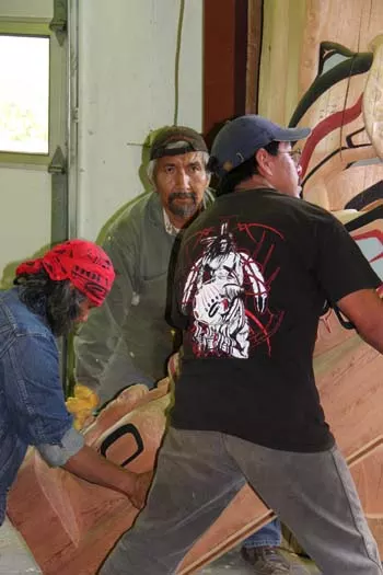 Three Indigenous men work together to lift a large, carved wooden house post