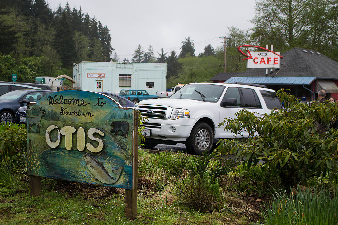a sign in the foreground reads "Welcome to downtown Otis". In the background we see rhododendrons, grasses and trees, a parking lot full of cars and SUVs and a sign for the Otis Cafe.